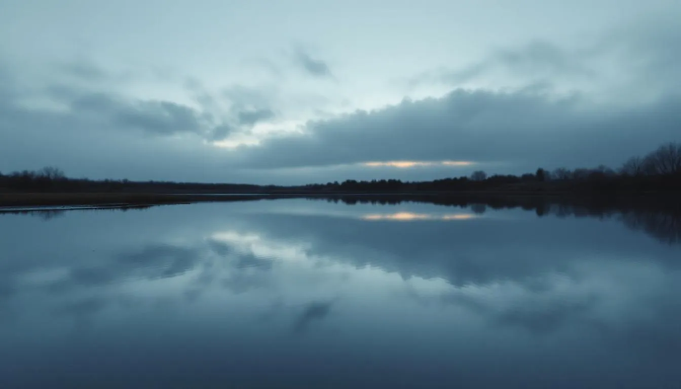 A calm lake at dusk mirrors a heavy grey sky with a faint break of orange light